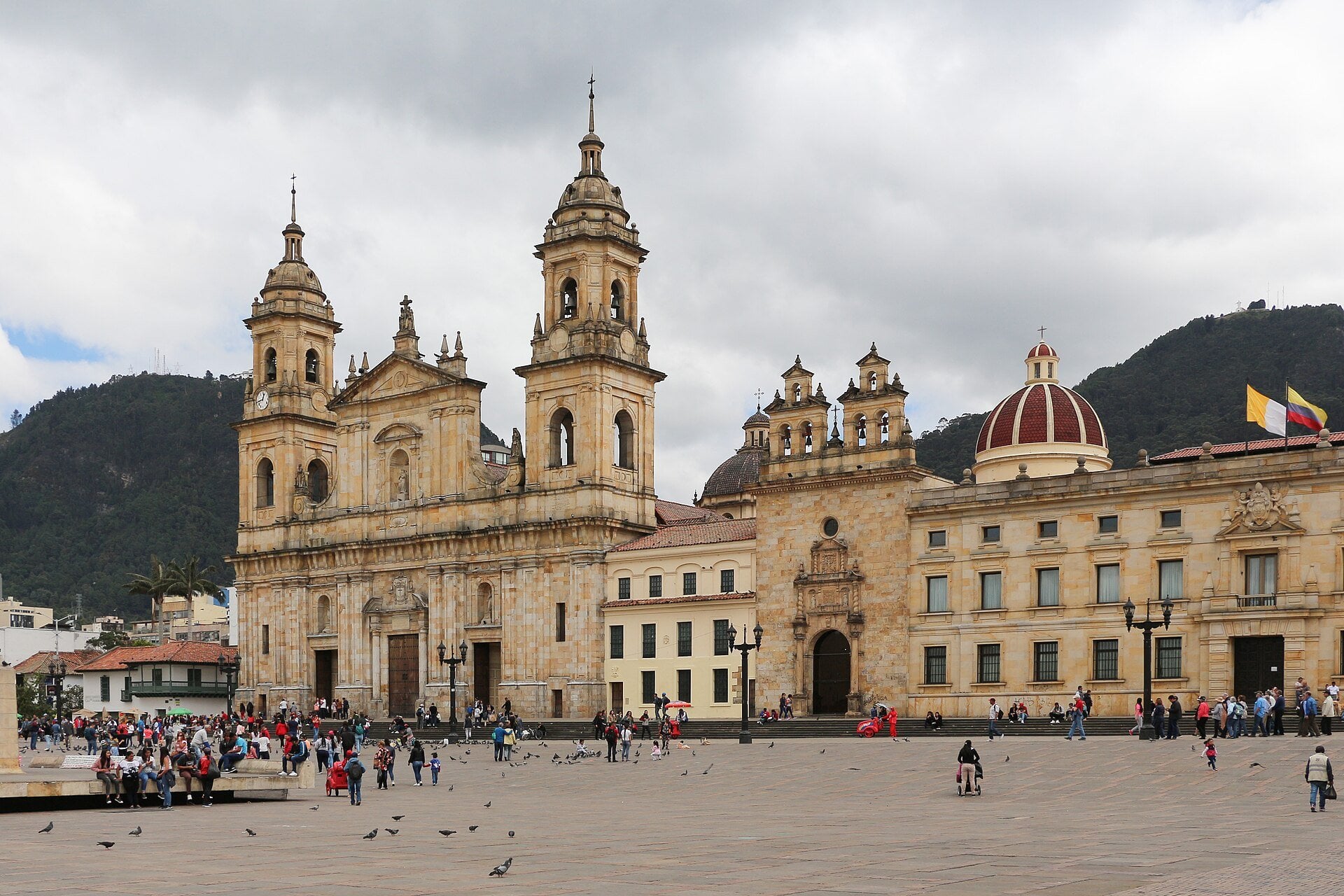 The Primate Cathedral of Colombia and neighboring colonial buildings on the Plaza de Bolívar in Bogotá, with people and pigeons on the square and green hills behind