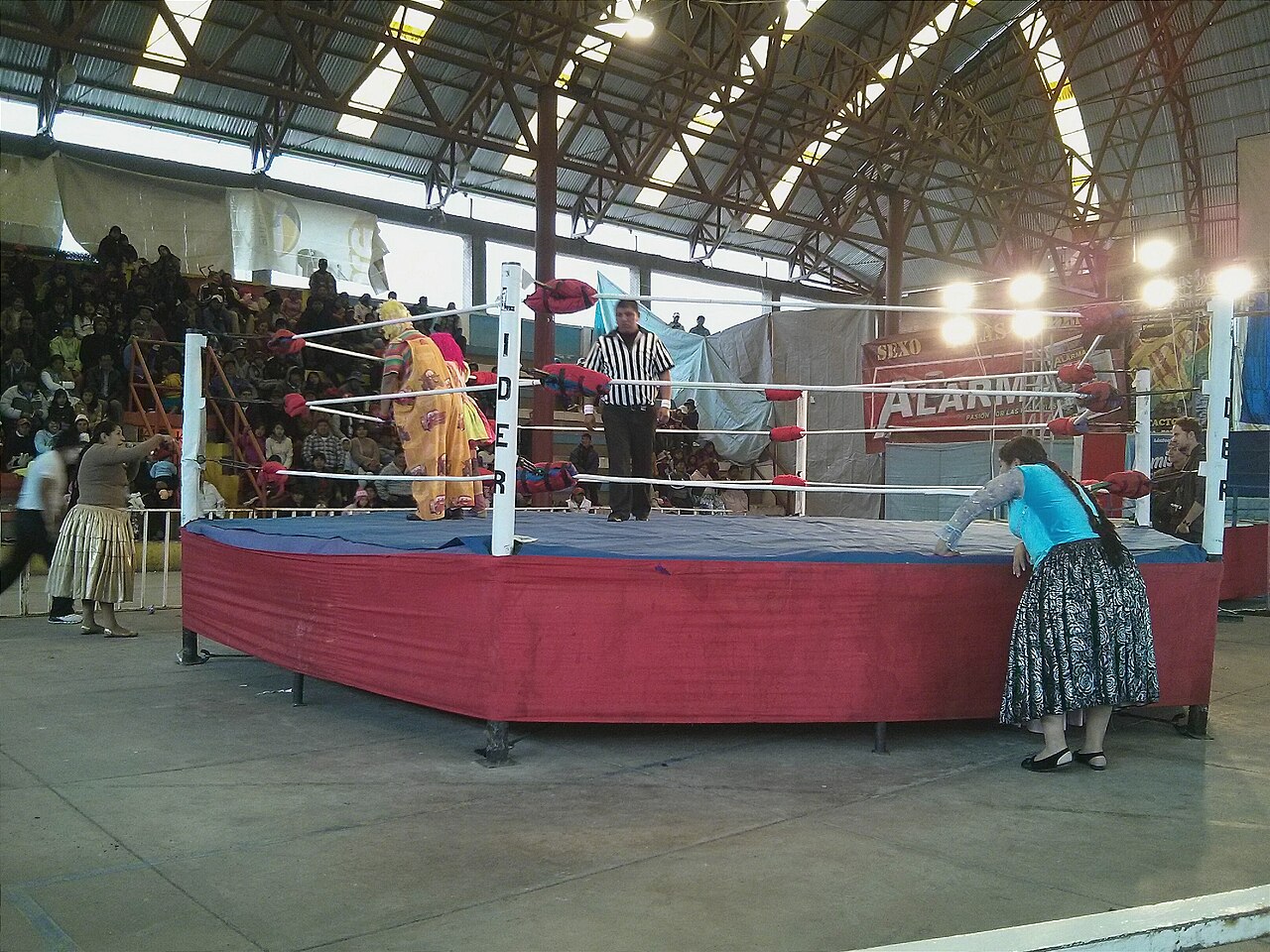 A wrestling ring in a covered hall in Bolivia with a referee and women in traditional polleras beside it