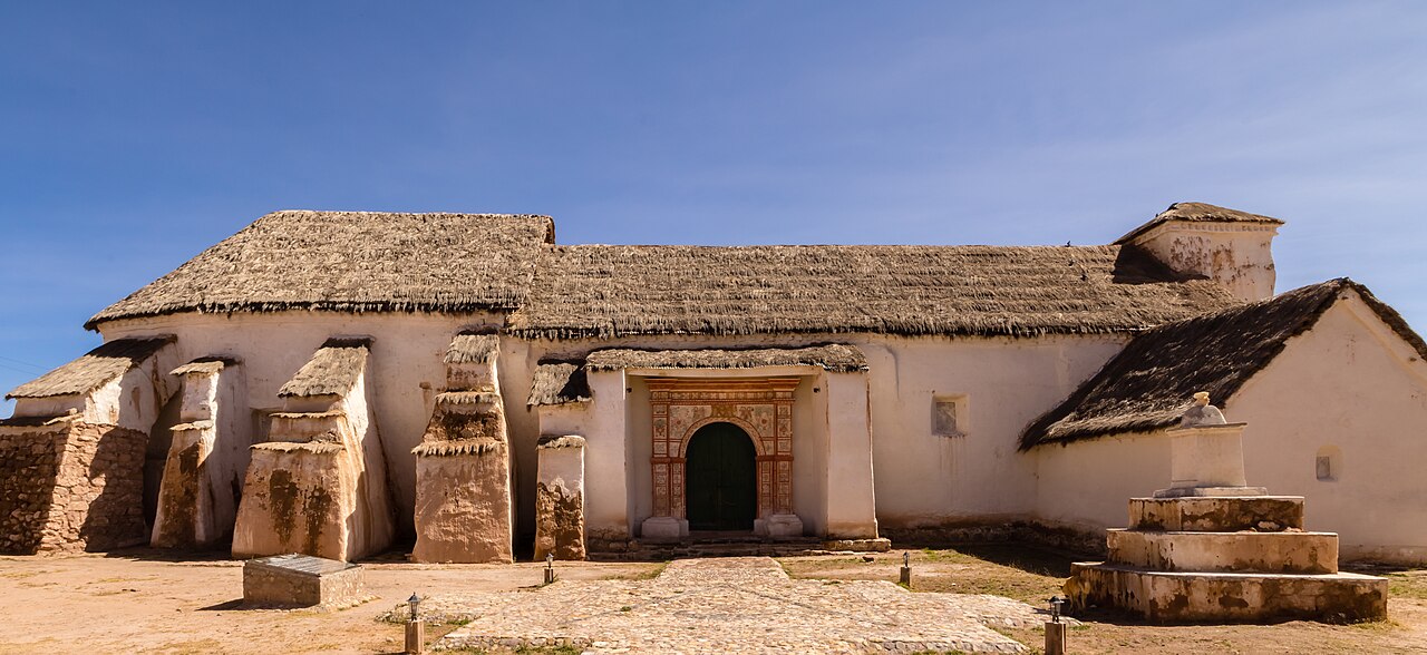 A whitewashed adobe church with thatched roofs, stone buttresses and a carved red-framed doorway under a clear blue sky, with a stone path in front