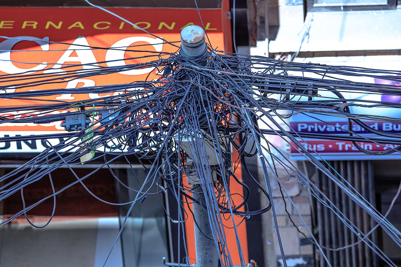 A dense tangle of black cables radiating from the top of a metal pole in front of shop signs in La Paz