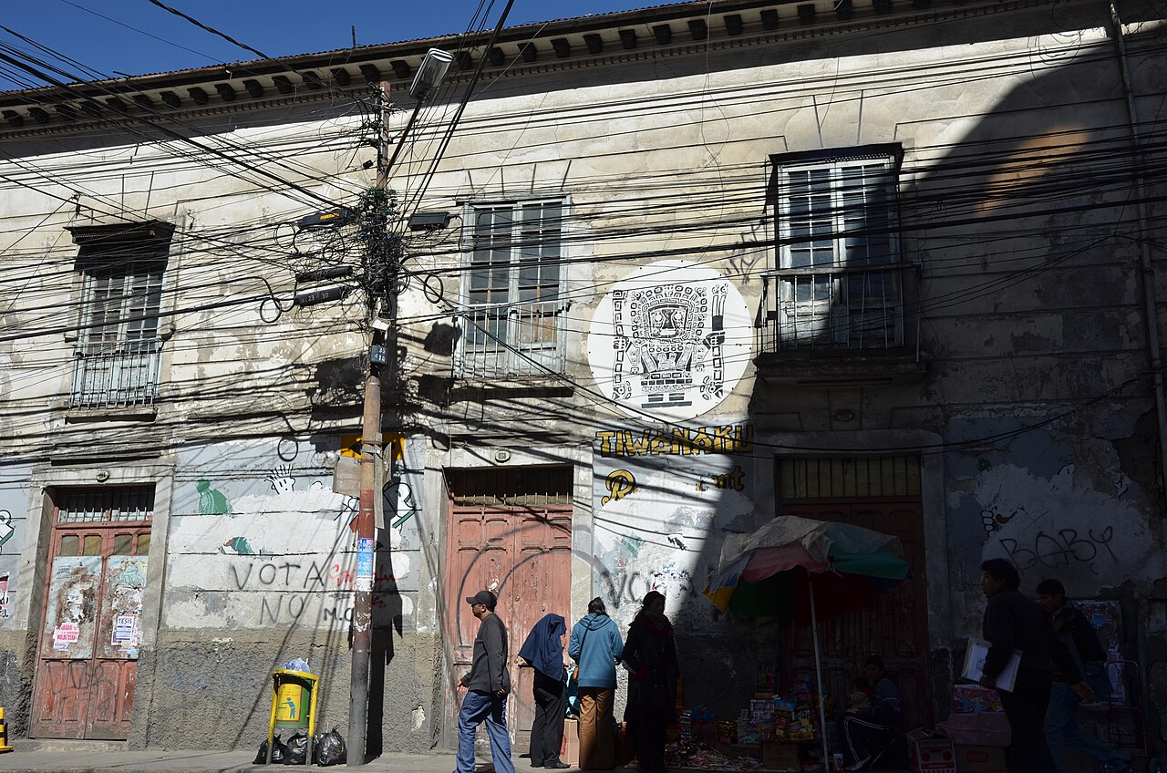 A colonial street in La Paz with dozens of overhead cables strung across a weathered facade, a Tiwanaku mural and pedestrians below