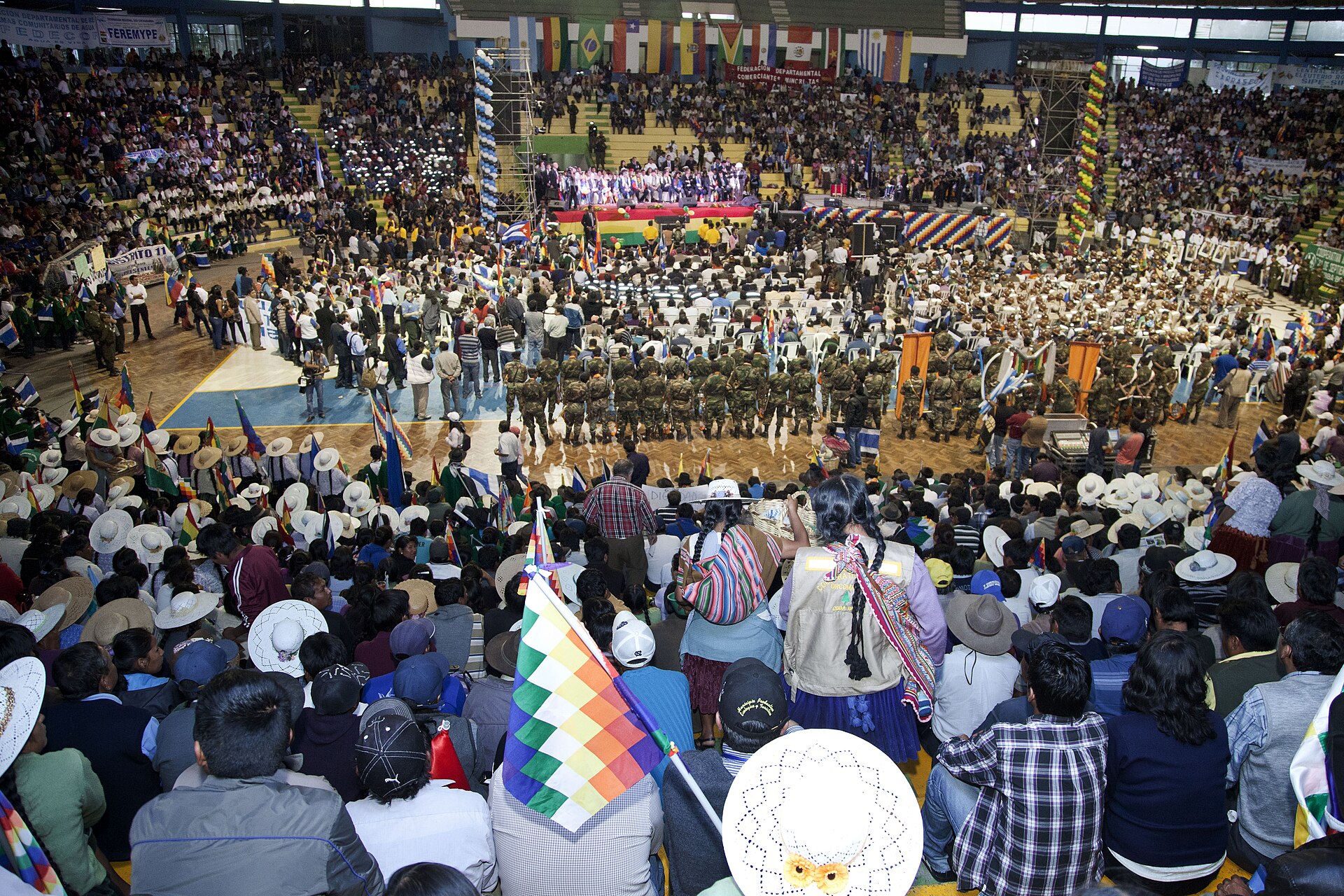 Packed indoor arena in Cochabamba with a stage at the far end, a crowd in hats and wiphala flags in the foreground and uniformed personnel standing in formation on the floor