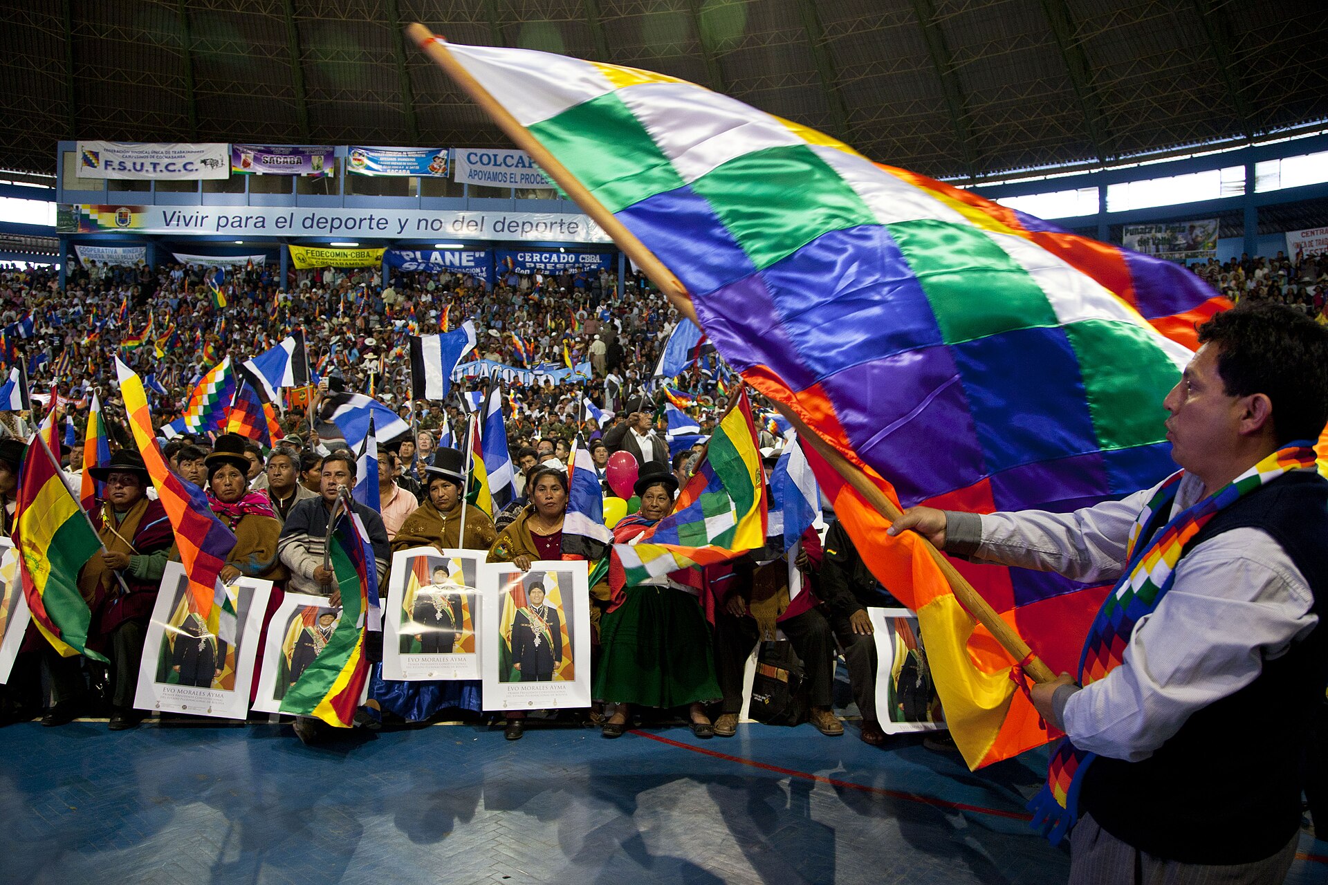 Crowd in a Cochabamba sports hall waves a large multicoloured wiphala flag beside supporters holding portraits of Evo Morales