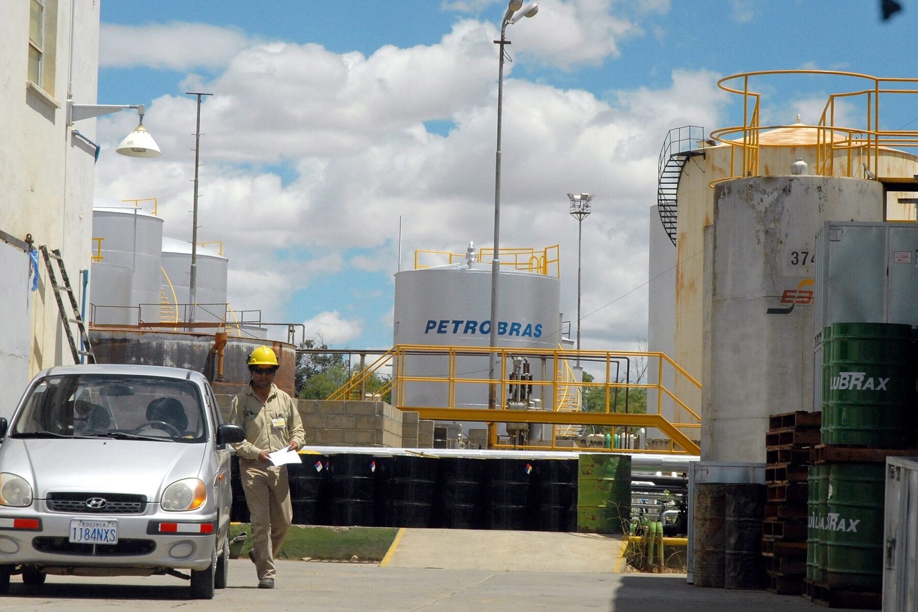 Worker in a yellow helmet walks past storage tanks marked Petrobras at a refinery in Cochabamba, Bolivia, with a silver car in the foreground