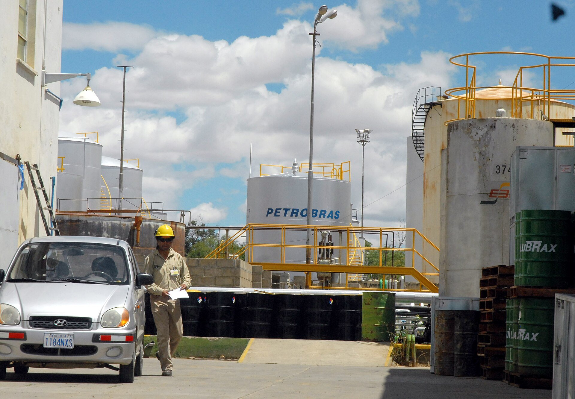 Worker in a yellow helmet walks past storage tanks marked Petrobras at a refinery in Cochabamba, Bolivia, with a silver car in the foreground
