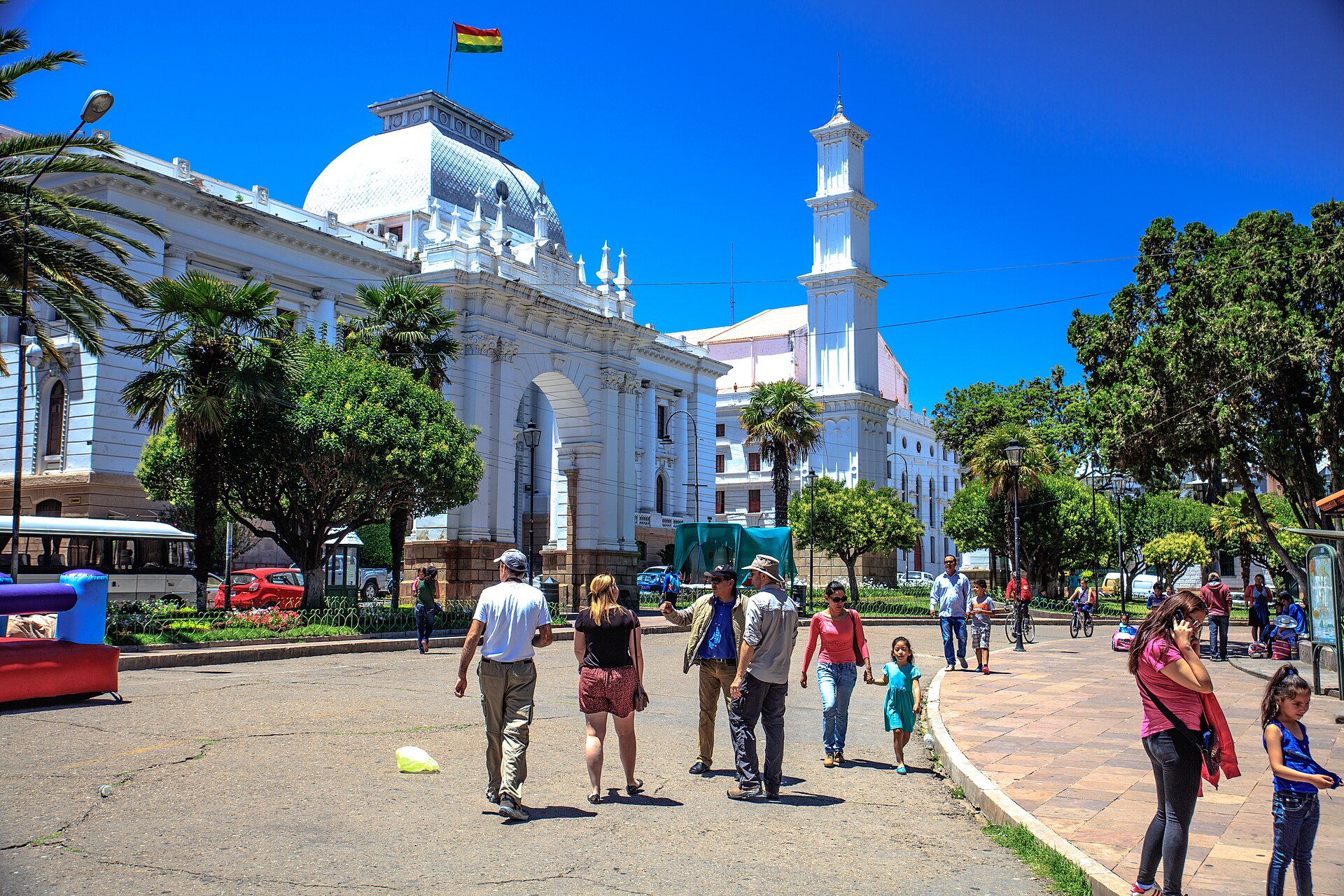 Pedestrians on a sunny plaza in Sucre, Bolivia, with a white domed palace, an arch and a bell tower behind
