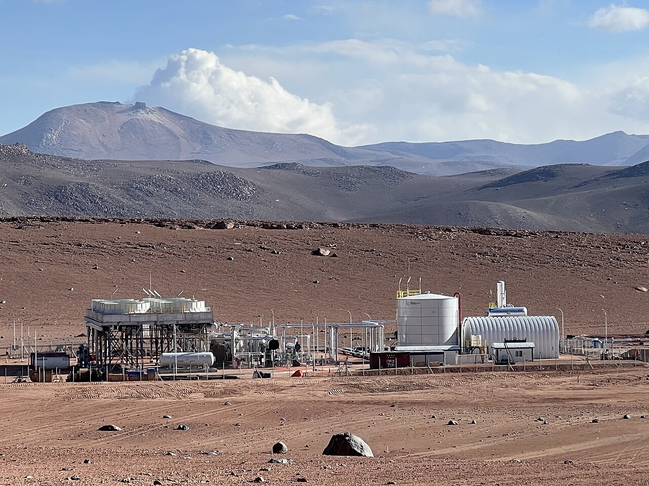 White tanks and pipework of a geothermal plant on a red desert plain below volcanic peaks
