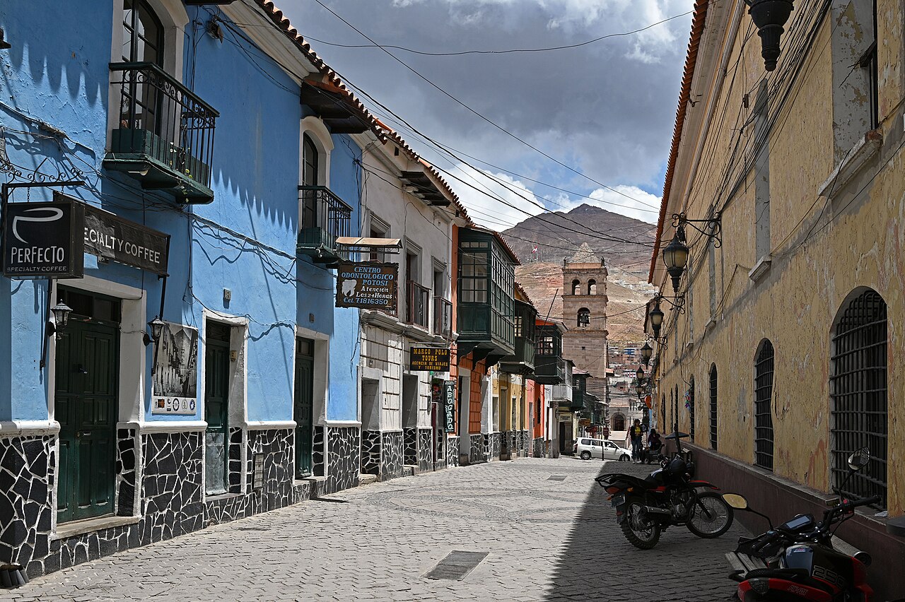 A cobbled colonial street in Potosí, Bolivia, with blue and white houses, wooden balconies, a church tower and the Cerro Rico mountain behind