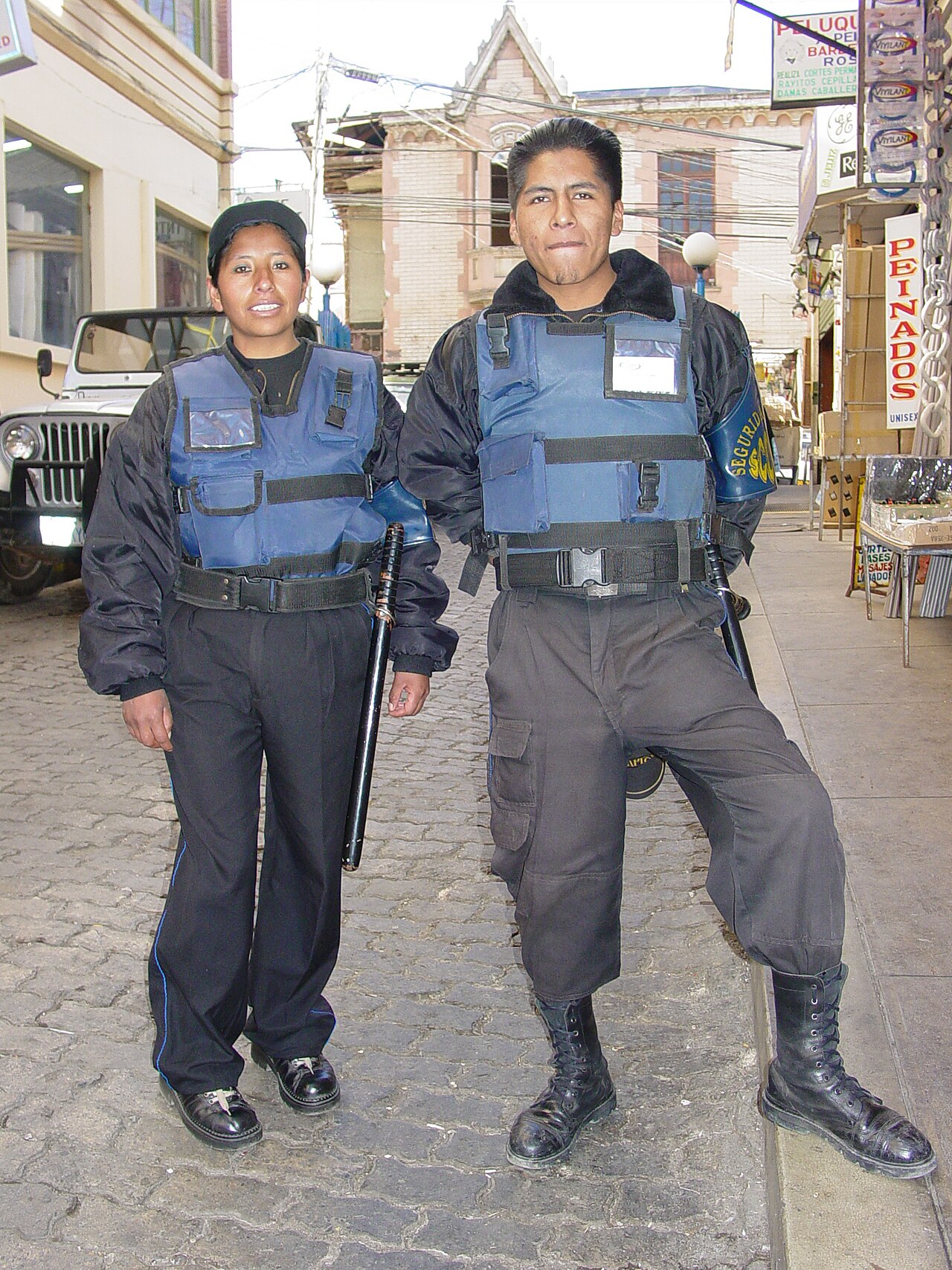 A female and a male officer in blue vests, carrying batons, stand on a cobbled street in La Paz.