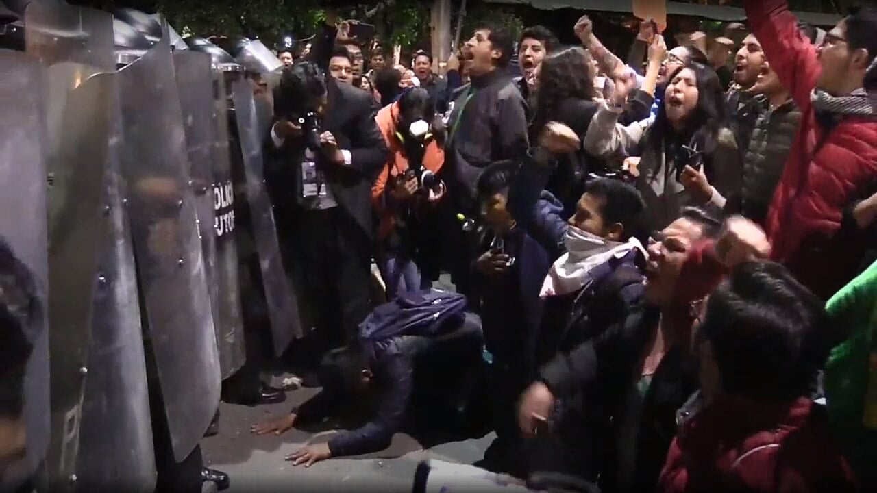 A row of metal riot shields marked Policía faces a crowd of shouting protesters at night in Bolivia.