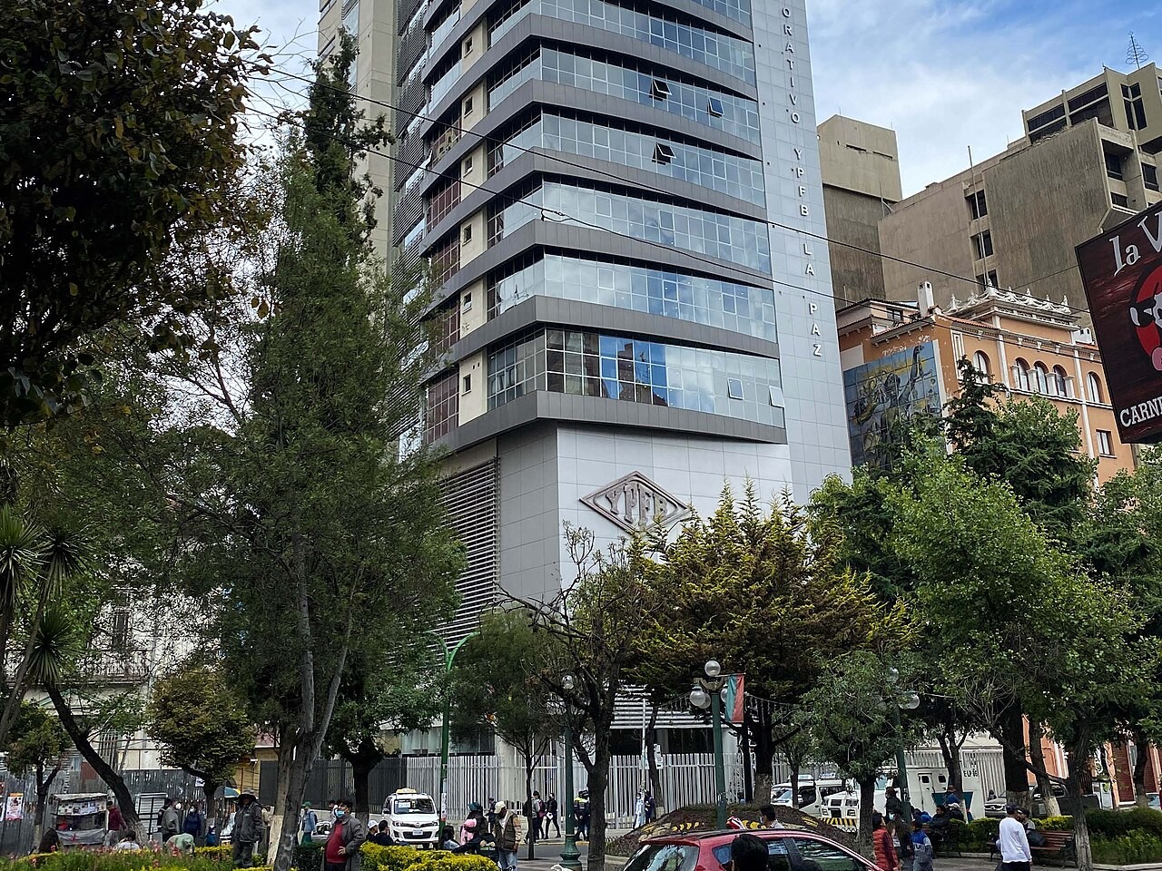 The glass-fronted YPFB corporate tower in La Paz with the YPFB logo on its facade, trees and pedestrians in a square in front