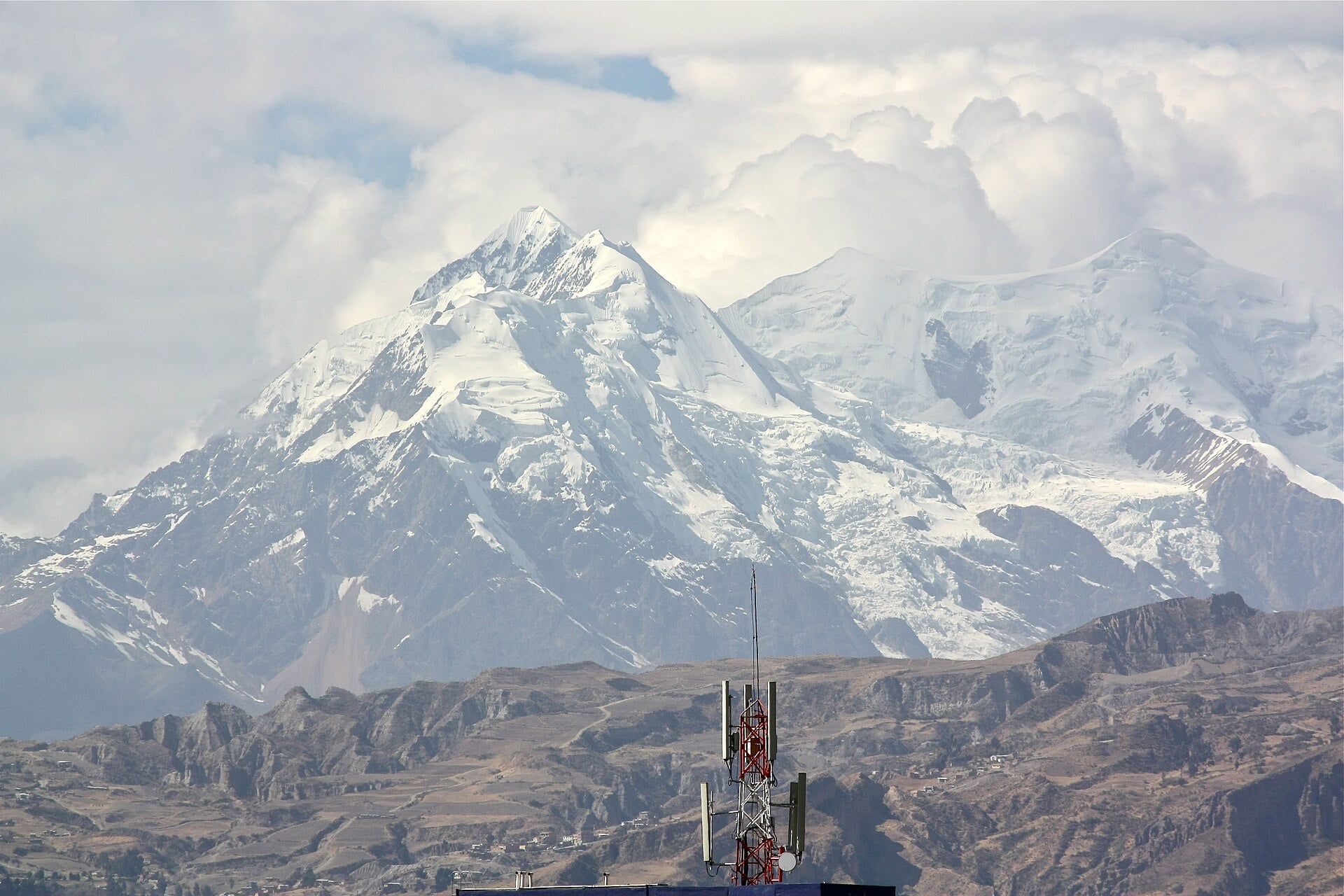 A red and white telecom mast on a hill in front of the snow-covered Illimani peak near La Paz