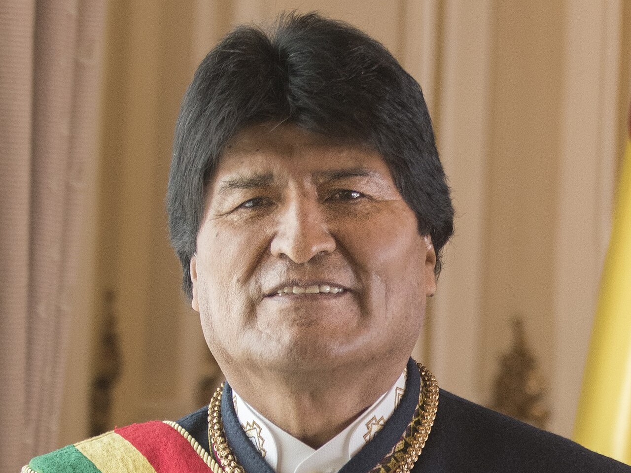 Evo Morales smiling in a formal portrait, wearing a dark jacket, gold chain and the red, yellow and green presidential sash of Bolivia