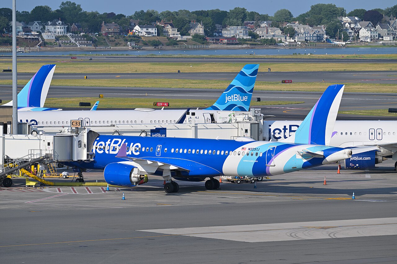 A blue-and-white JetBlue jet parked at a gate at Boston Logan airport, with other jets and waterfront houses behind