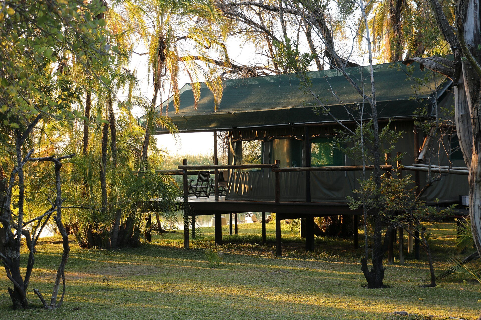 A green canvas safari lodge on a raised wooden deck among palm trees beside the water in the Okavango Delta, Botswana