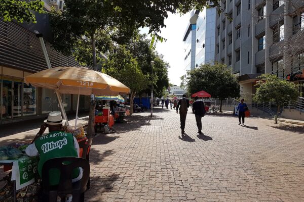 Pedestrians walk along a tree-lined paved mall in central Gaborone, with a street vendor under an umbrella and office buildings on the right