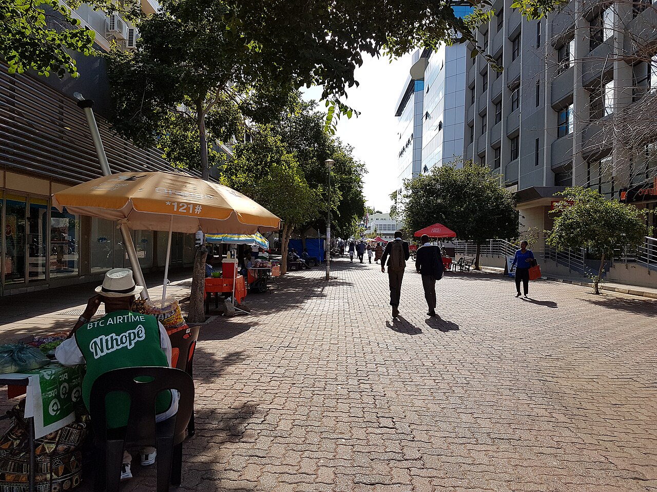 Pedestrians walk along a tree-lined paved mall in central Gaborone, with a street vendor under an umbrella and office buildings on the right
