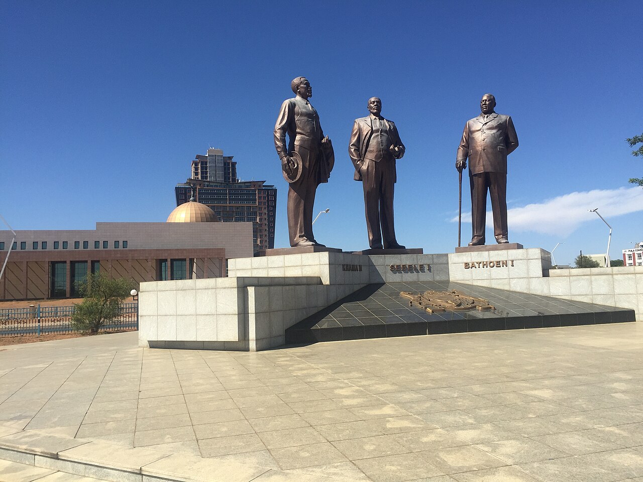 Three bronze statues of Botswana chiefs on a stone platform in front of government buildings