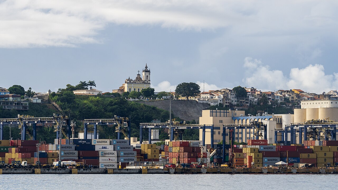 Stacks of shipping containers and blue gantry cranes at a port terminal, with a hillside church and houses behind