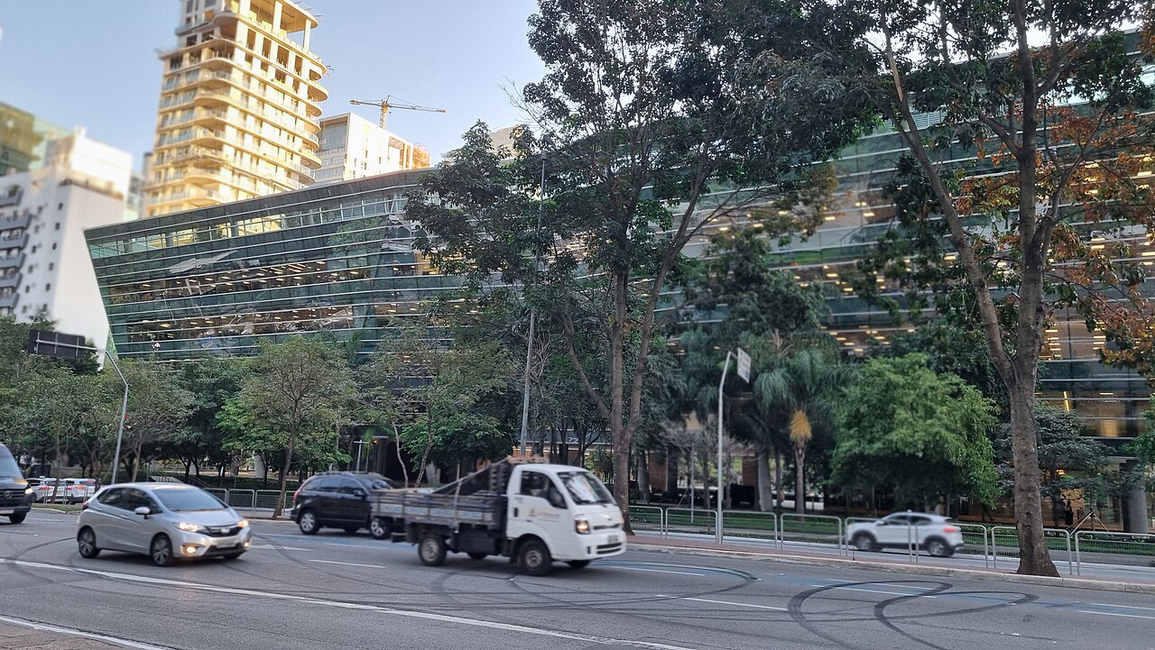 Cars and a small truck pass a glass office building and towers on Avenida Faria Lima in São Paulo