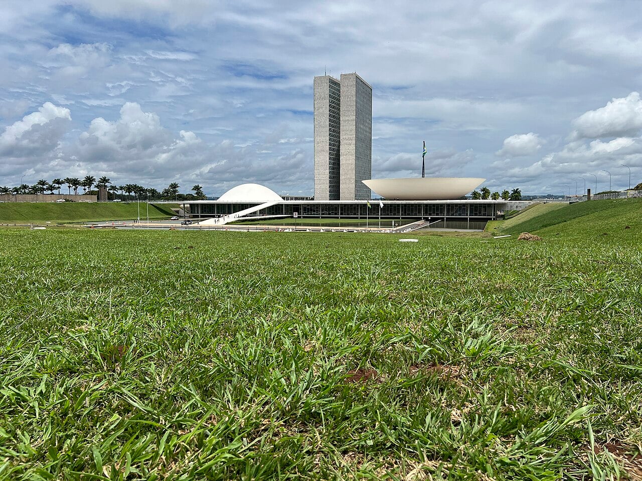 The National Congress building in Brasília, with its twin towers, white dome and bowl, seen across a green lawn under a cloudy sky