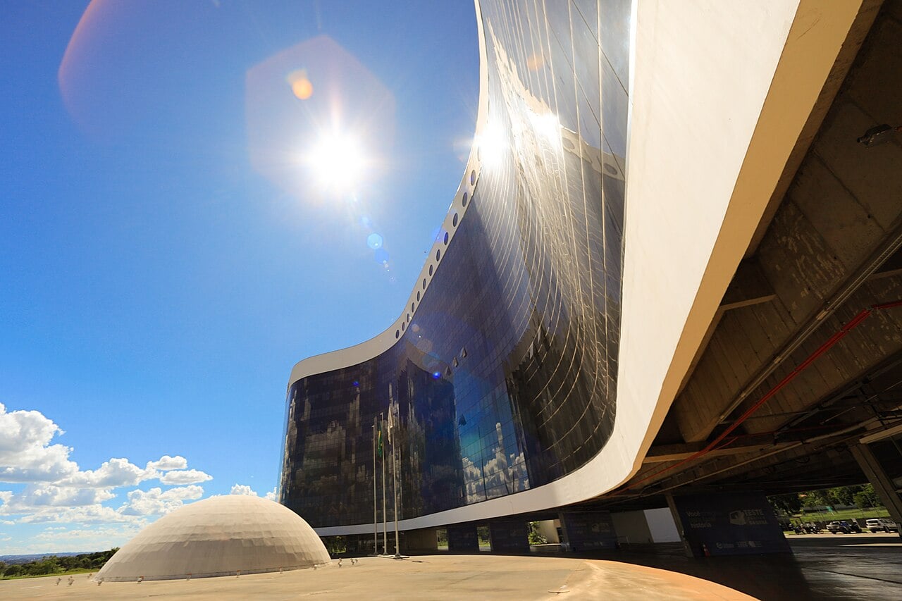 Curved dark glass tower and a white dome of the Superior Electoral Court headquarters in Brasília under a bright sun and blue sky