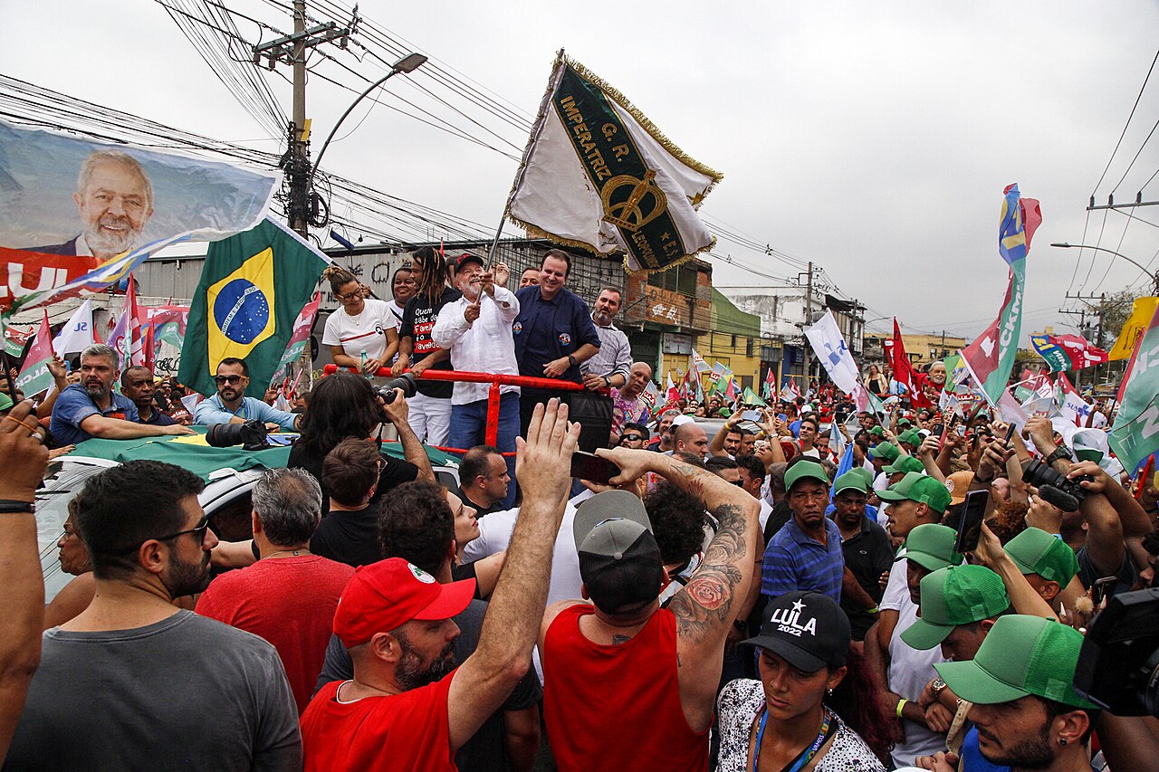 Lula waves a samba school flag from the top of a vehicle as supporters in red and green caps crowd the street during a car rally