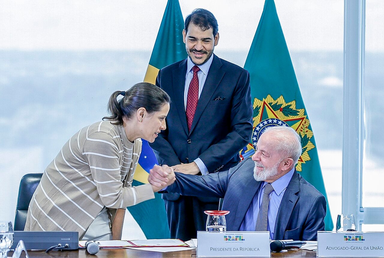 President Lula, seated, shakes hands with Pernambuco governor Raquel Lyra as Jorge Messias stands behind them between two Brazilian flags