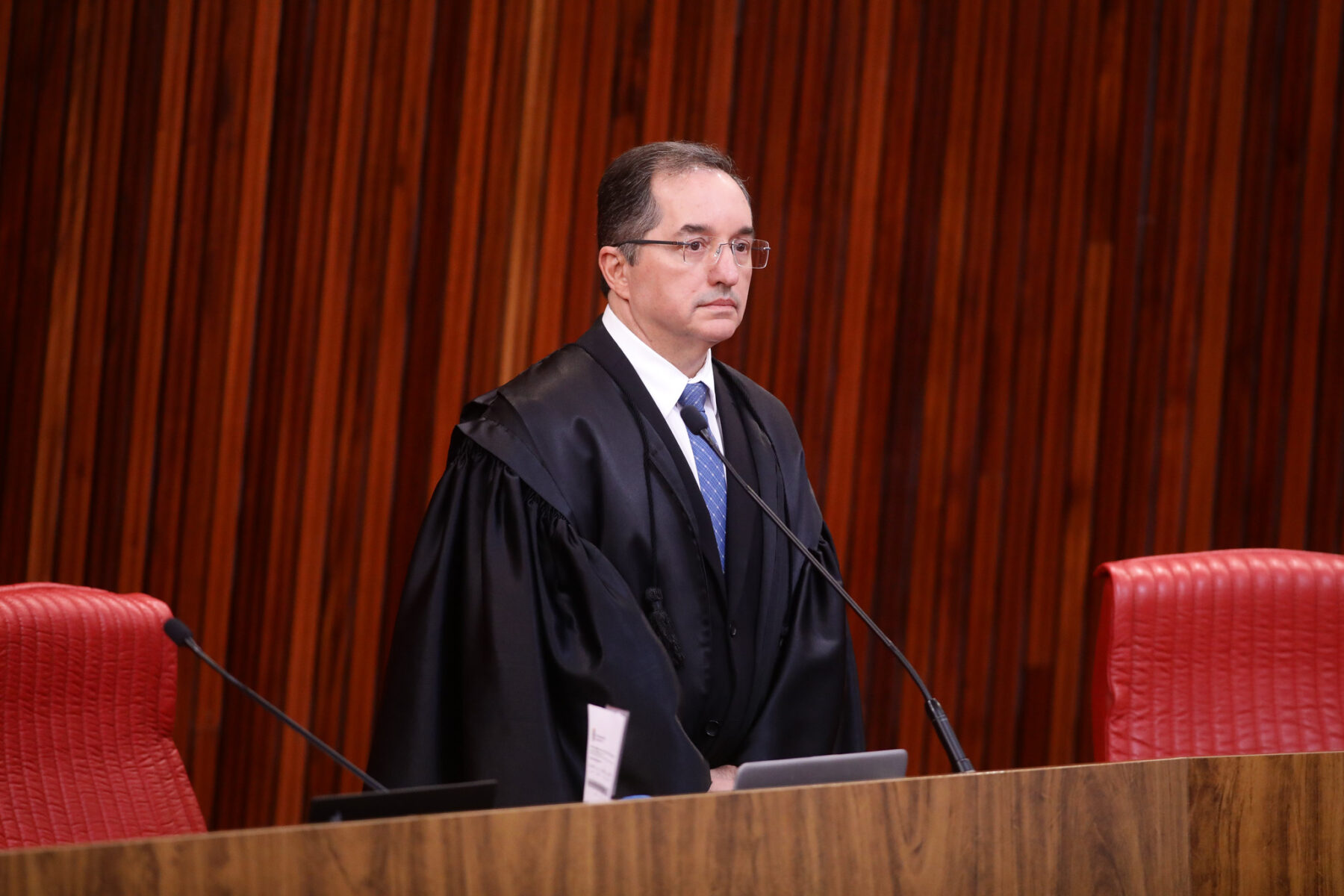 A judge in black robes standing at the bench of the Superior Electoral Court (TSE) plenary in Brasília, with wood-panelled walls and red leather chairs