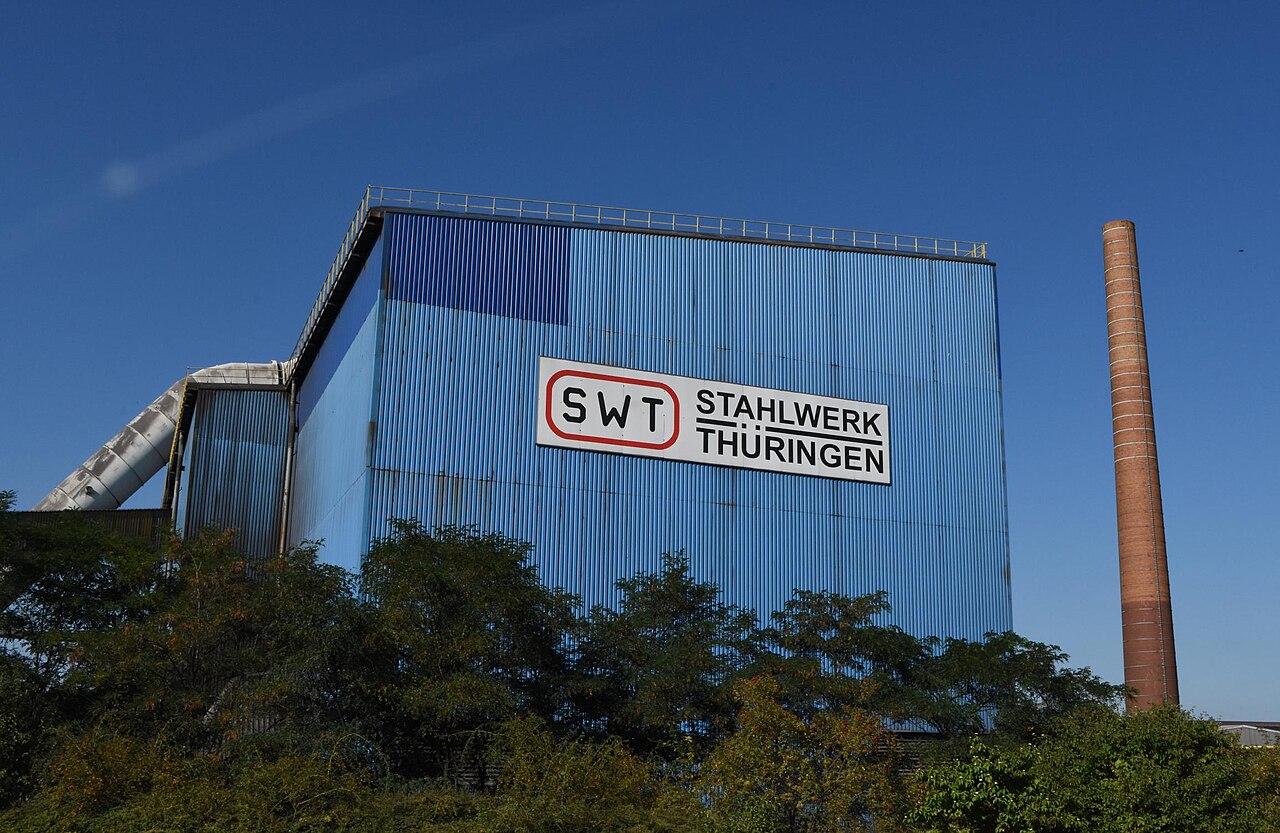 A tall blue steel-clad mill building with a white Stahlwerk Thüringen SWT sign and a brick chimney beside it, trees in the foreground