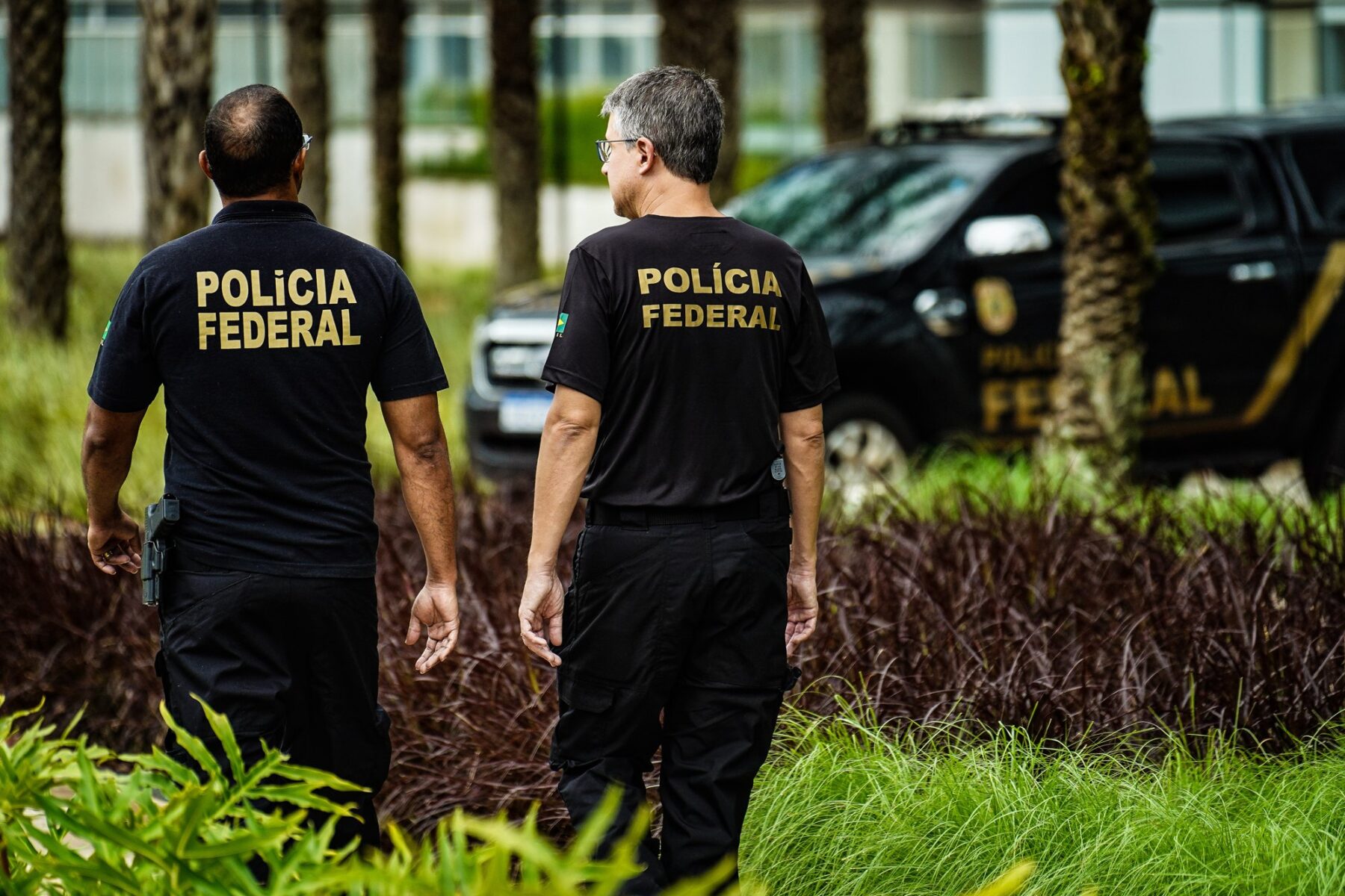 Two Brazilian Federal Police officers in black uniforms walk past a marked Federal Police vehicle in Brasília