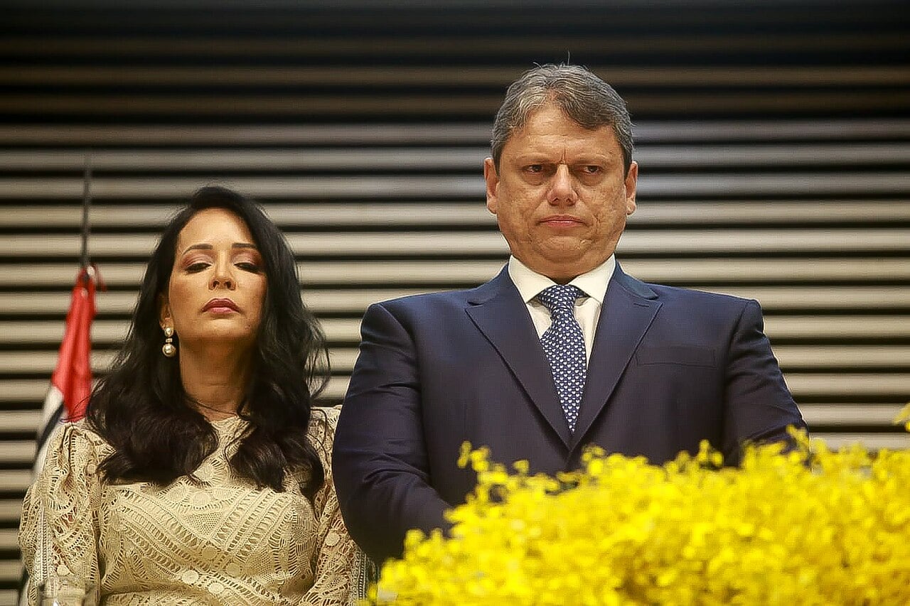 Tarcísio de Freitas, in a dark suit and blue patterned tie, stands at a ceremony with a woman beside him and yellow flowers in the foreground