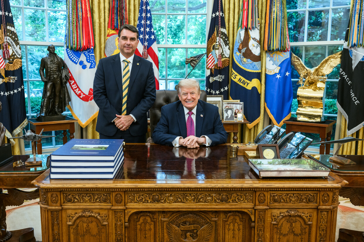 Flávio Bolsonaro stands beside Donald Trump, who sits smiling at the Resolute desk in the Oval Office