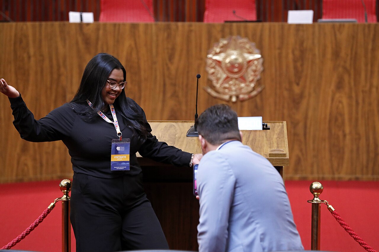 Visitors stand beside the speaker's lectern in the wood-panelled plenary chamber of Brazil's Superior Electoral Court, with the national coat of arms behind and rope barriers in front