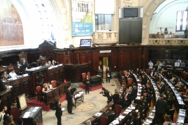 A session in the wood-panelled plenary chamber of the Rio de Janeiro state assembly, with participants at curved desks and visitors in the gallery