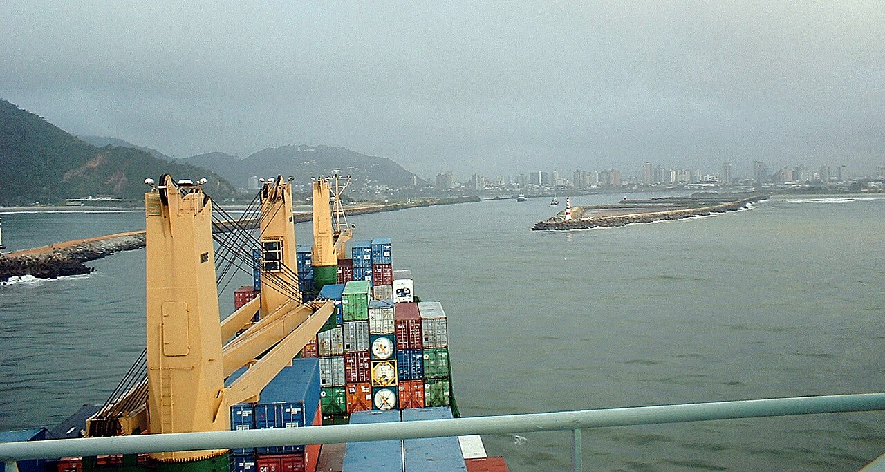 Stacked shipping containers on a cargo ship entering the harbour at Itajaí, Brazil, between two stone breakwaters with the city skyline behind