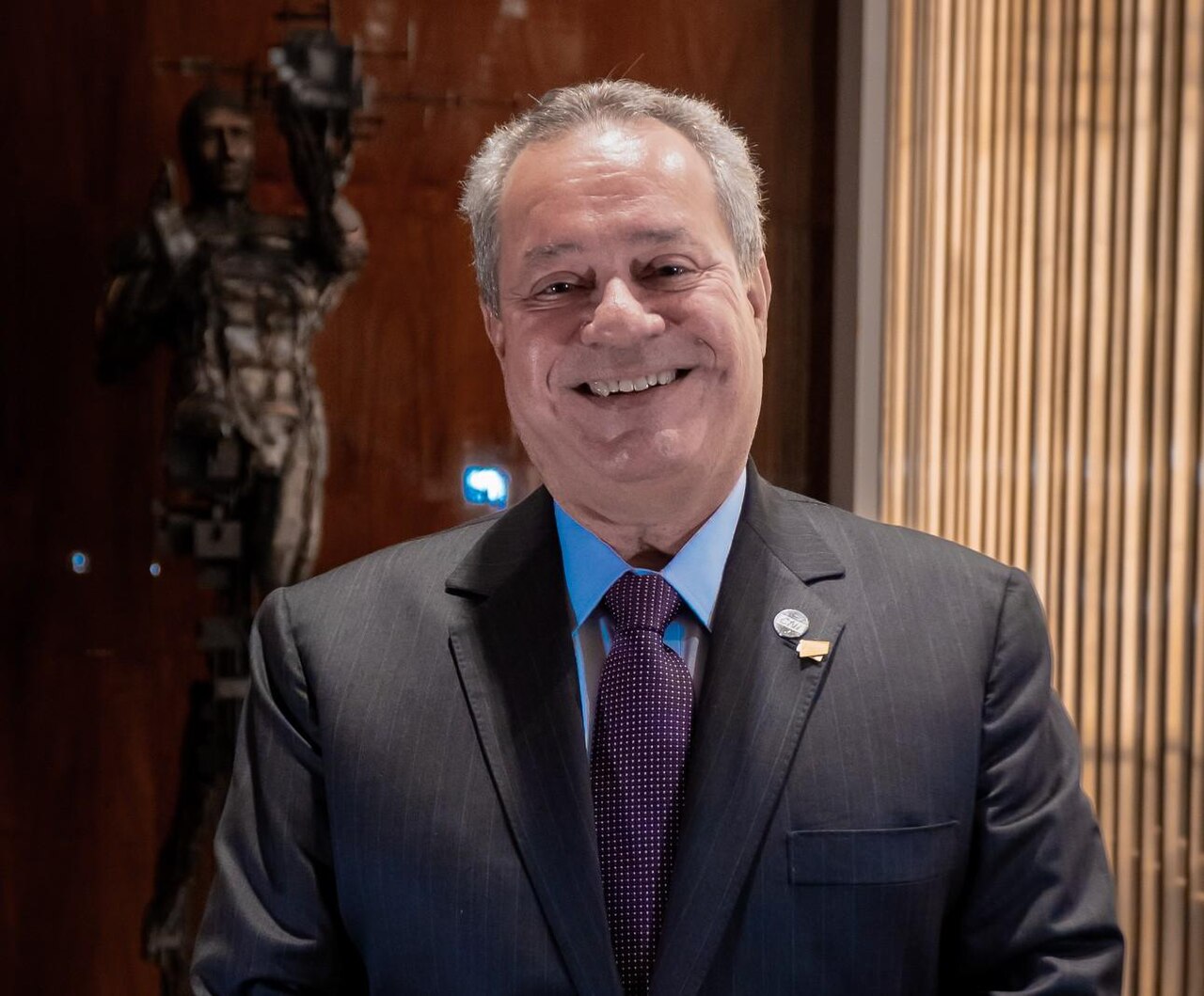 Ricardo Alban, president of the National Confederation of Industry, smiles in a dark suit and purple tie with a lapel pin, in a wood-panelled room