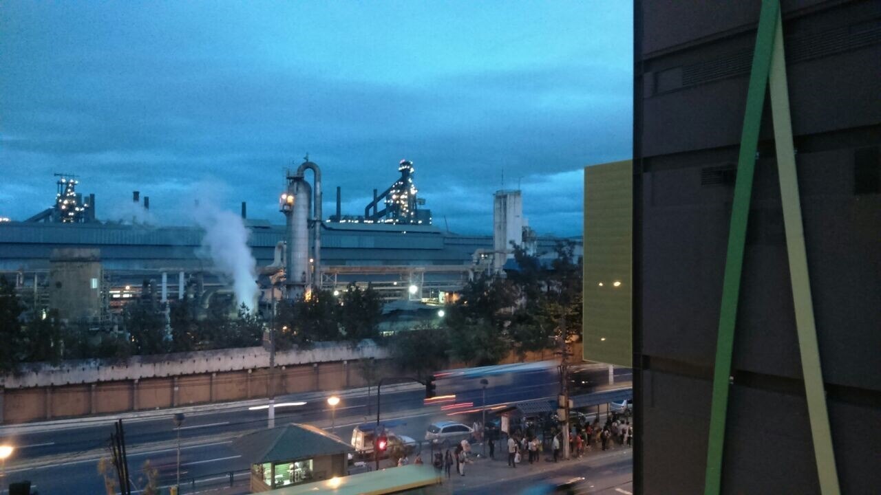 Blast furnaces and a steam plume at the CSN steelworks in Volta Redonda, Brazil, at dusk, seen across a busy road with a bus stop
