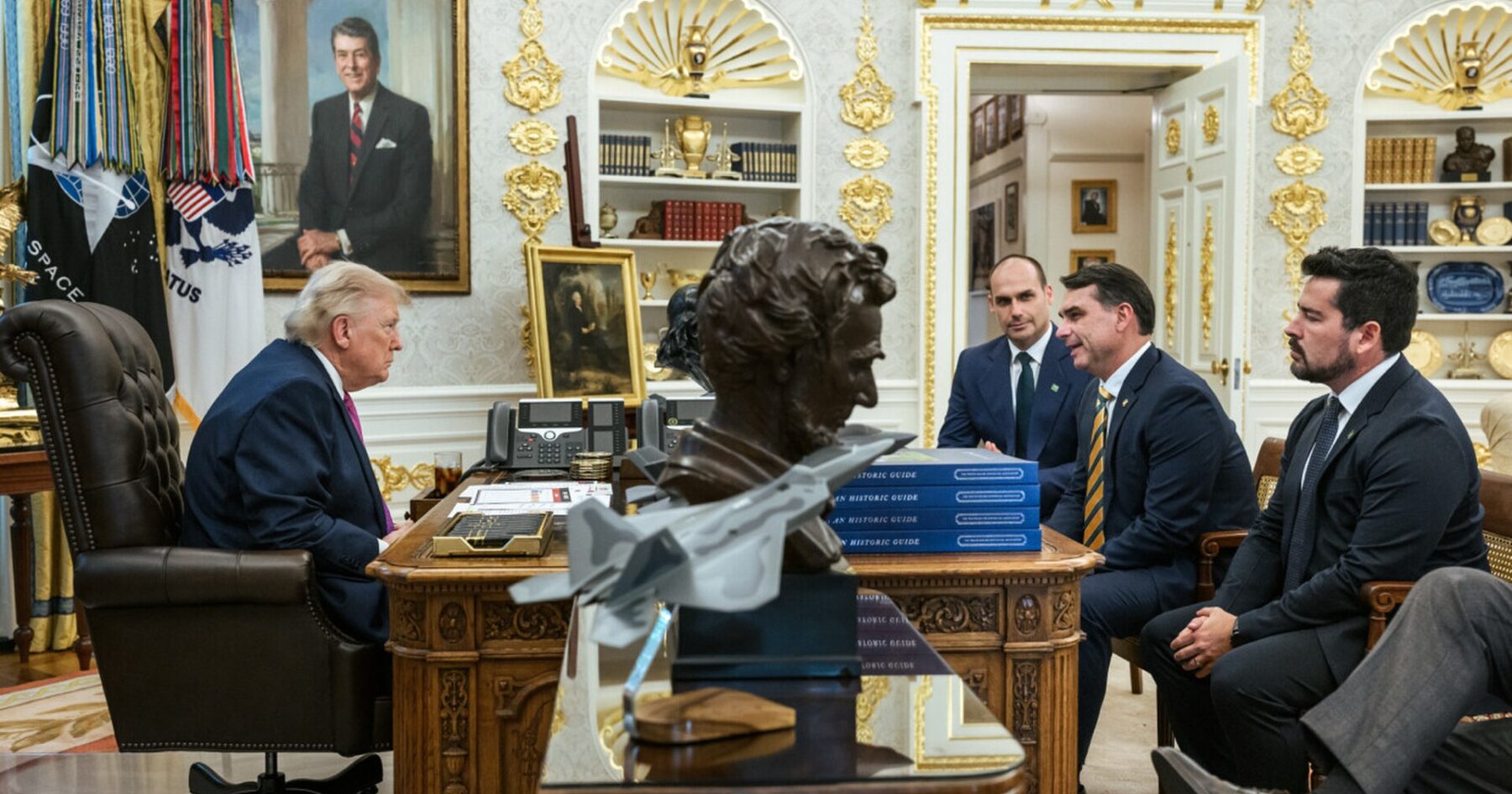 President Donald Trump seated at his desk in the Oval Office, with Brazilian senator Flávio Bolsonaro and two other men seated opposite