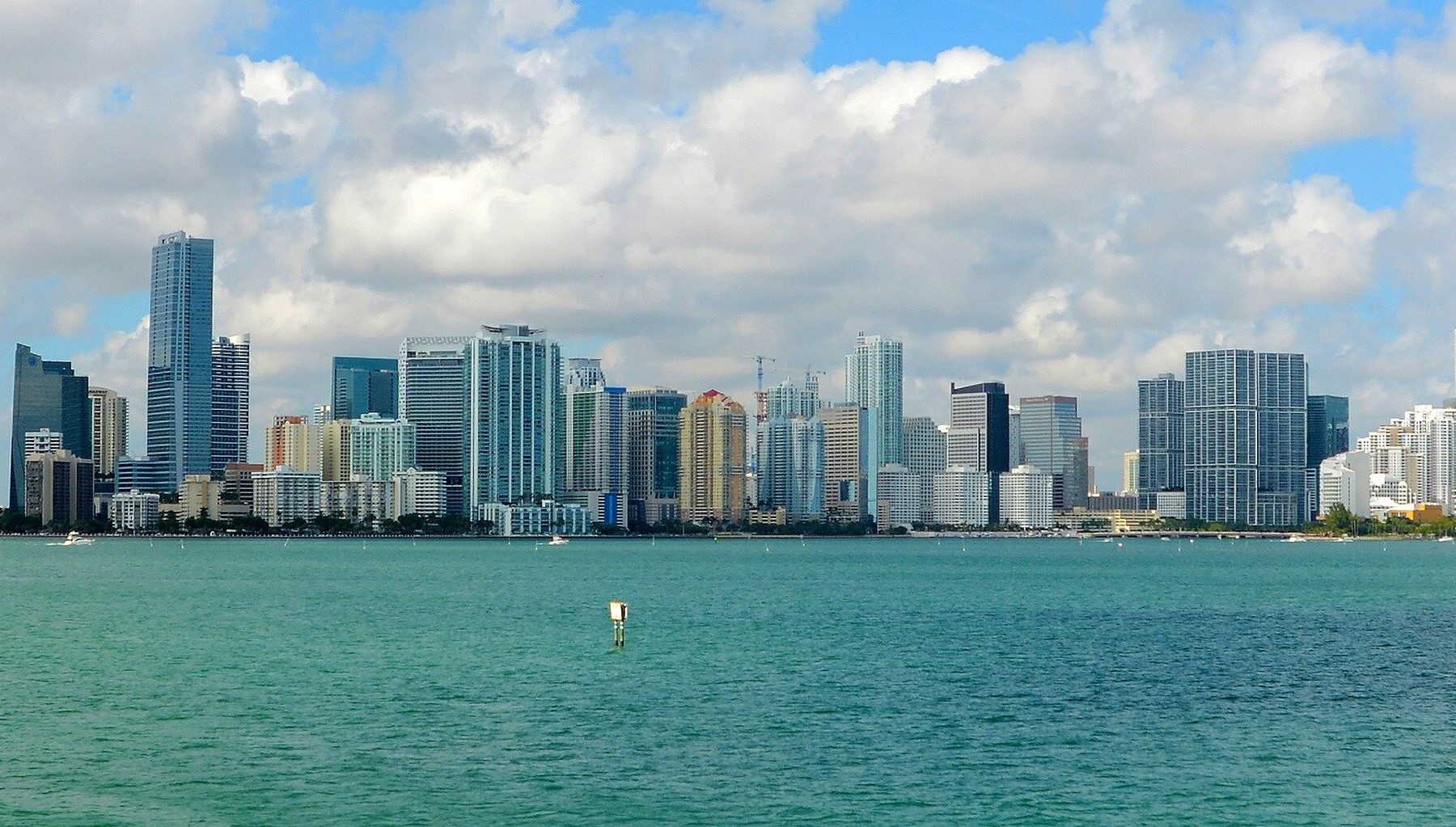 The Miami skyline with tall glass towers seen across the blue-green water of Biscayne Bay under a cloudy sky