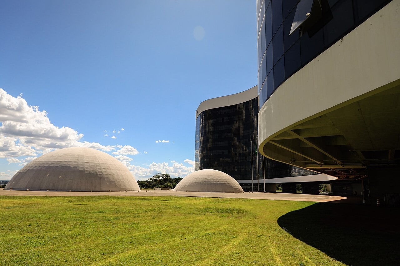 Two white domes and the curved dark glass tower of the Superior Electoral Court headquarters in Brasília under a blue sky