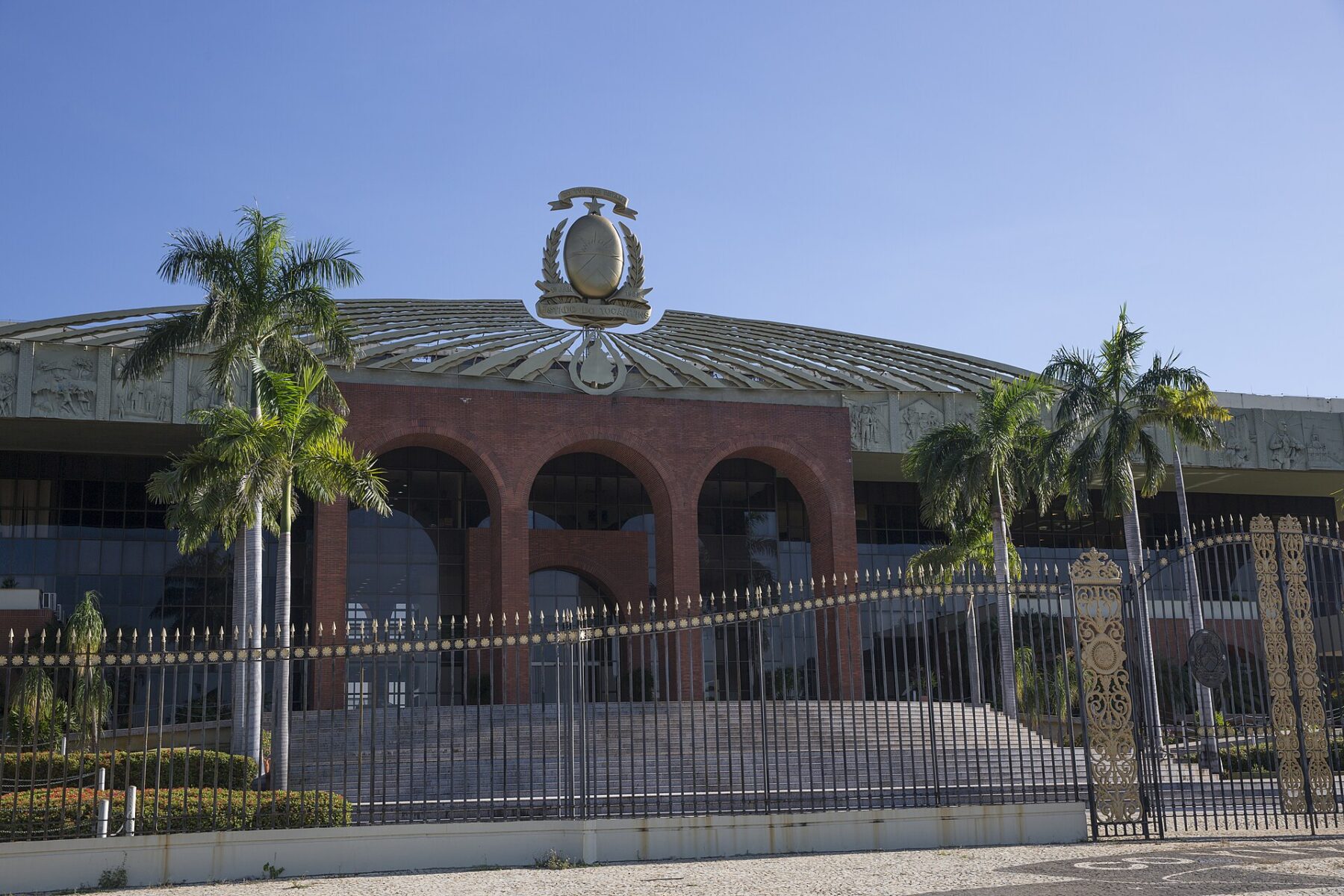 The brick arches and gilded coat of arms of the Palácio Araguaia government palace in Palmas, Tocantins, behind palm trees and an iron fence