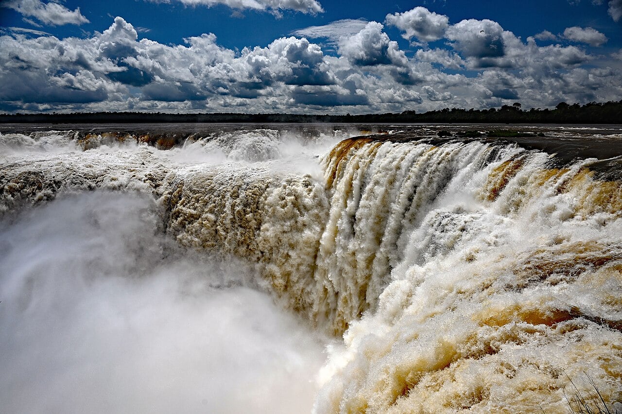 Brown floodwater pours over the curved rim of the Devil's Throat at the Iguaçu Falls under a blue sky with white clouds