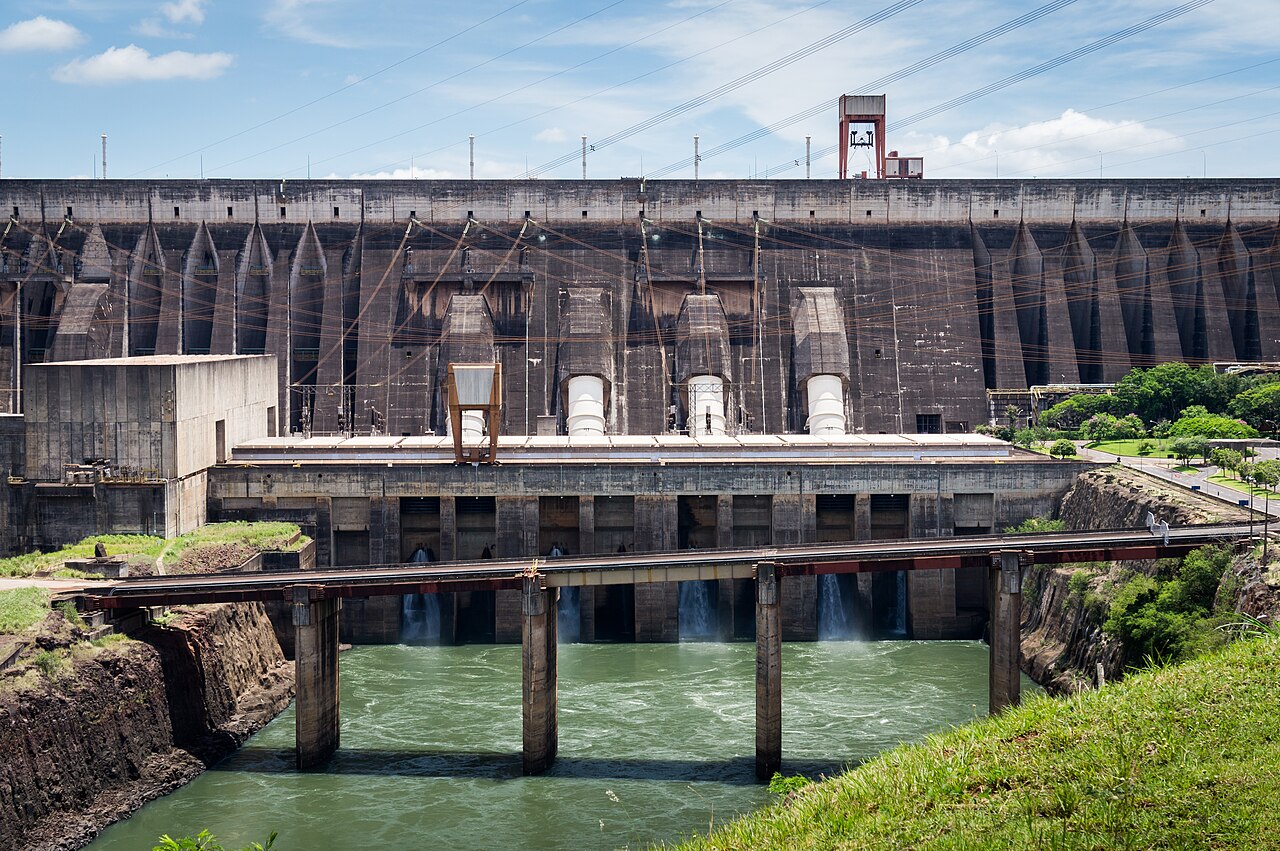 Concrete face of the Itaipú dam with water released from outlets below the powerhouse into a green river channel crossed by a low bridge