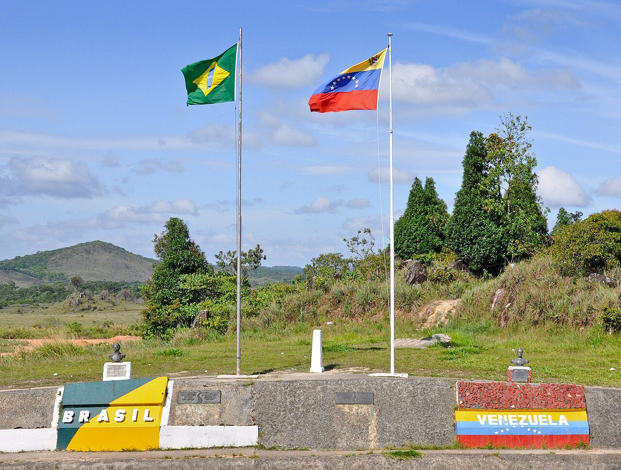 Brazilian and Venezuelan flags fly over a border marker with painted Brasil and Venezuela signs and two small busts in green hilly country