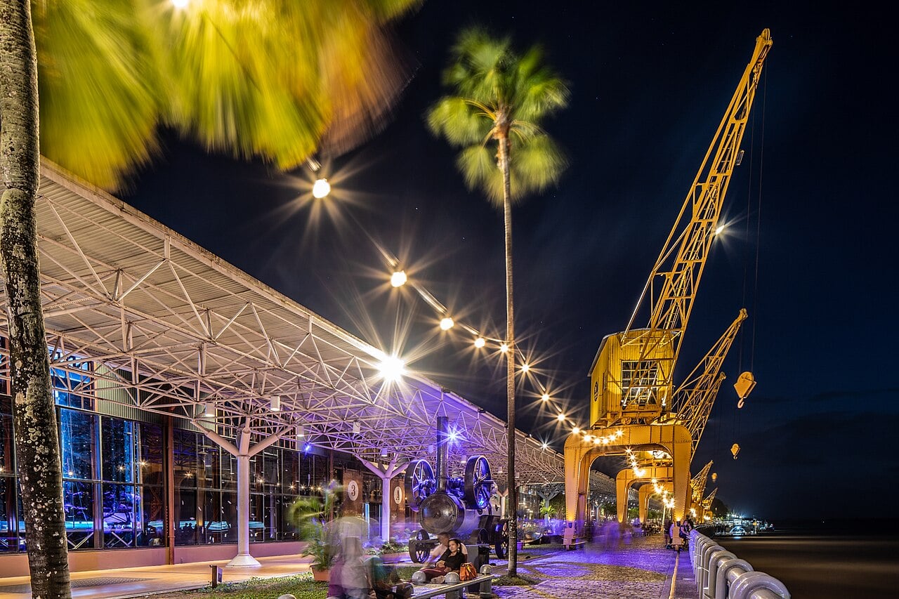 Riverside promenade at night in Belém with a lit steel-roofed warehouse, palm trees, string lights and yellow port cranes