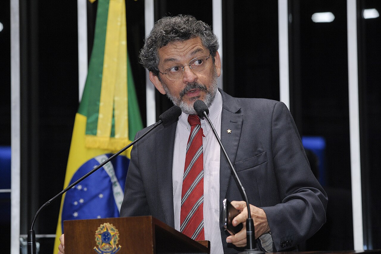 A grey-haired man with glasses and a red striped tie speaks at a lectern in front of a Brazilian flag