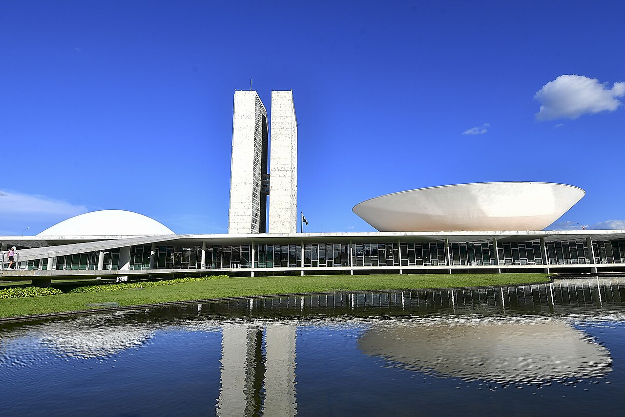 The twin towers, white dome and upturned bowl of the National Congress building in Brasília, reflected in a pool under a blue sky