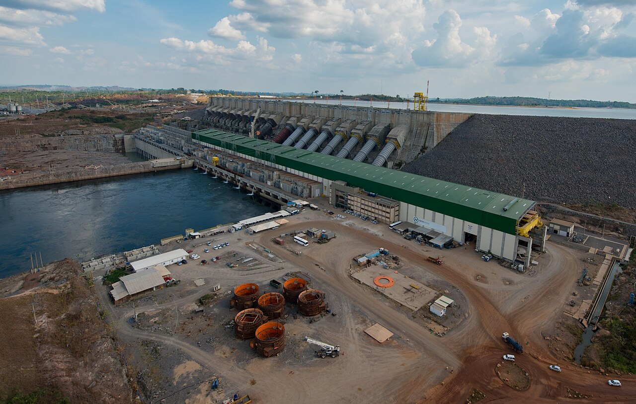 Aerial view of a hydroelectric powerhouse with a green roof, large penstocks and a reservoir behind a dam