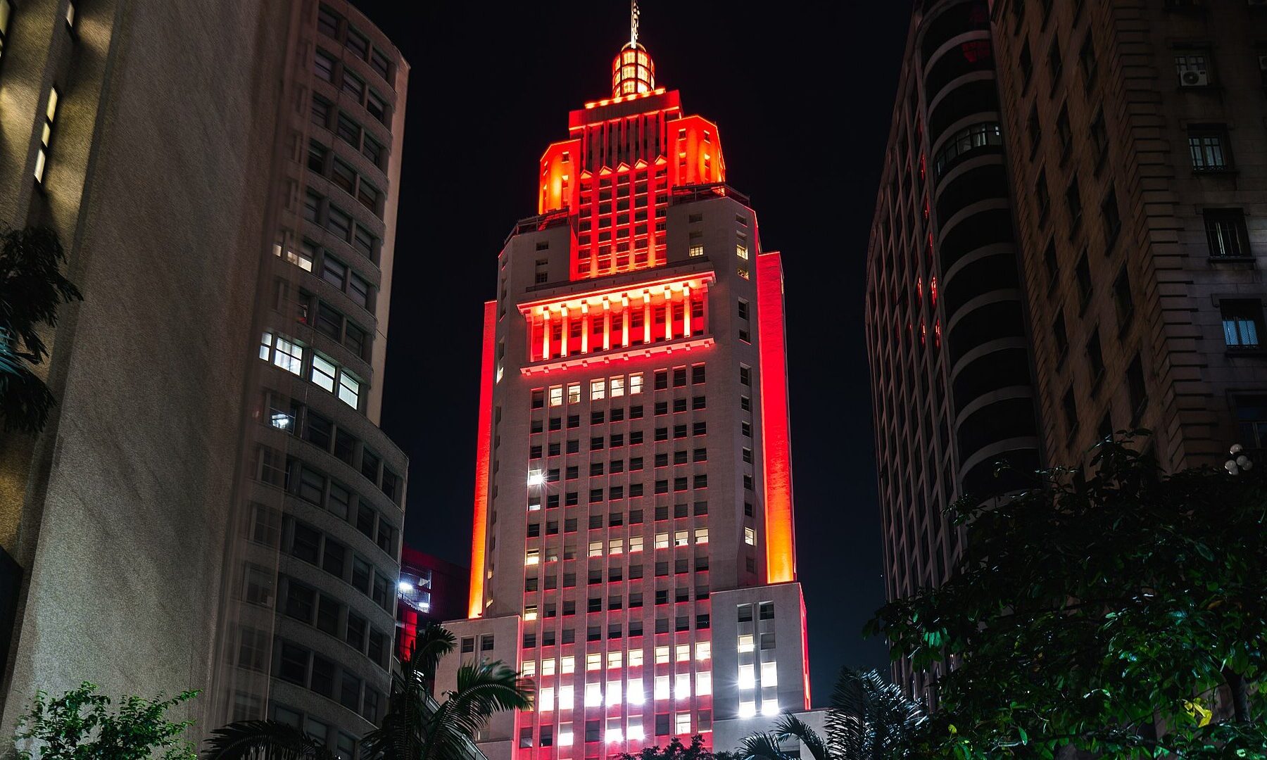 The Altino Arantes skyscraper in downtown Sao Paulo lit in red and orange at night between neighbouring buildings and palm fronds